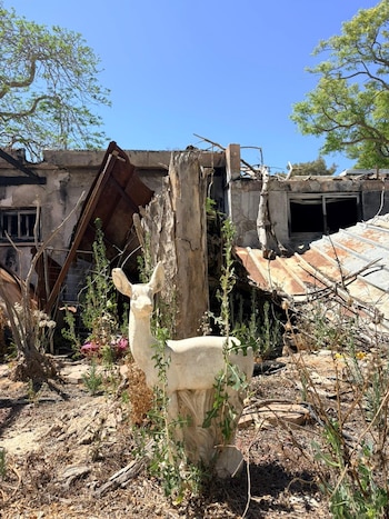 Vista exterior de una estatua de ciervo de piedra en primer plano, con un edificio destruido y escombros cubiertos de vegetación en el fondo bajo un cielo azul claro