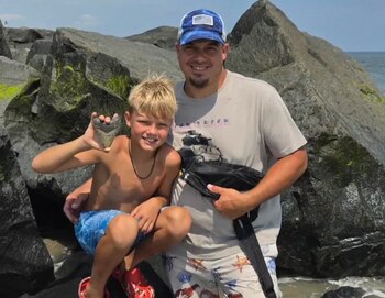 Un hombre y un niño posan sonriendo junto a rocas en la orilla del mar, el niño sostiene un diente grande de color claro