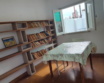Una habitación con estantes de madera y bloques de hormigón llenos de libros, una mesa cubierta con un mantel y una ventana abierta