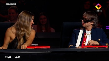 Jimena Barón, con cabello rubio y blusa negra brillante, mira a Abel Pintos, quien usa chaqueta azul militar, camiseta blanca, cadena dorada y gafas de sol