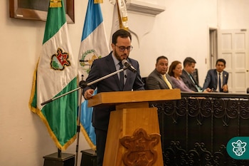 Hombre con traje y gafas hablando en un atril con micrófono, con banderas de Guatemala y otra verde y blanca detrás, y personas sentadas en el fondo