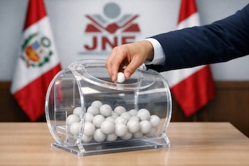 Mano sacando una bolilla blanca de un ánfora transparente llena de bolillas. Al fondo, banderas de Perú y el logo desenfocado del JNE en una mesa de madera.