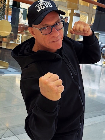 Jean-Claude Van Damme con gorra negra, gafas y sudadera deportiva levanta ambos puños cerrados frente a la cámara en un espacio interior