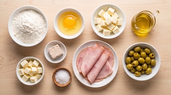 Recipientes con harina, un huevo, cubos de queso, aceite, levadura, sal, jamón en fetas y aceitunas verdes dispuestos sobre madera.