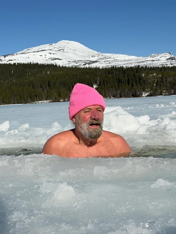 Hombre con barba y gorro rosa sumergido hasta el pecho en un lago helado con trozos de hielo, con montañas nevadas y un bosque de pinos al fondo