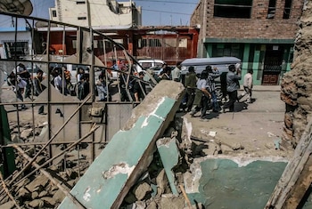 Varias personas cargan un ataúd por una calle con escombros. Se ven edificios dañados, una reja metálica rota y niños detrás de ella