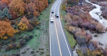 Vista aérea de tres vehículos y personas sobre una carretera de dos carriles rodeada de árboles otoñales y un curso de agua