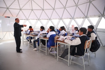 Un profesor de traje gesticula frente a estudiantes uniformados sentados en pupitres, dentro de un aula en forma de cúpula geodésica blanca con pizarra