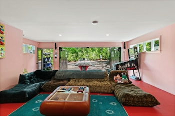 Sala de juegos moderna con paredes rosas y suelo rojo. Incluye sofás, mesas de futbolín y pinball, y acceso a un exterior verde con un carrito de golf azul