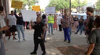 Varias personas protestan en una acera, algunas sostienen pancartas con mensajes como 'Silence is Violence' y 'Chief Fraser Do Your Job' frente a edificios