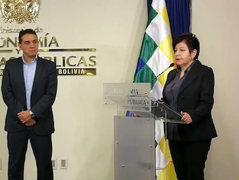 Una mujer habla ante un micrófono en un atril transparente junto a un hombre de traje frente a la bandera wiphala y un muro institucional