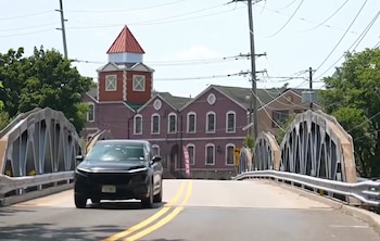 Un vehículo oscuro transita por un puente de metal con dos carriles y marcas amarillas. Al fondo se ven edificios de ladrillo y torres, con un cielo claro