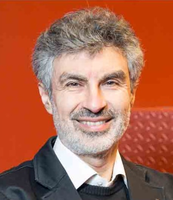 Retrato de Yoshua Bengio, un hombre de mediana edad con cabello gris, barba y bigote, sonriendo directamente a la cámara frente a un fondo rojo