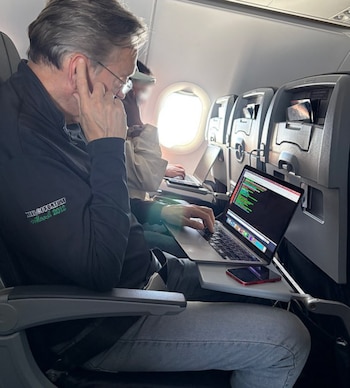 Un hombre con gafas sentado en un avión utiliza una computadora portátil sobre la mesa plegable de su asiento