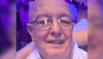 Primer plano de un hombre calvo, de piel clara y gafas, con una camisa blanca, sonriendo ligeramente. El fondo está desenfocado con tonos púrpuras y azules