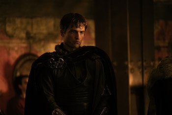 Robert Pattinson, medio cuerpo, viste túnica oscura con capa de piel y broches. Fondo interior con iluminación tenue. Hombro de otra persona visible a la derecha