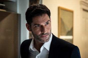 Primer plano de Tom Ellis, un hombre de cabello oscuro y barba, vistiendo un traje oscuro y camisa blanca, mirando fijamente a su derecha. Fondo interior borroso