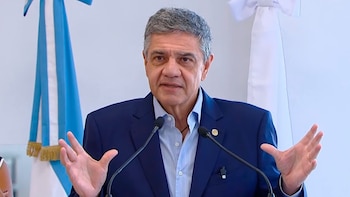 Primer plano de Jorge Macri, un hombre con cabello gris y traje azul, hablando en un podio con dos micrófonos, con la bandera argentina de fondo