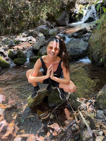 Mujer en cuclillas sobre rocas dentro de un arroyo, con ropa deportiva negra y gafas de sol, manos unidas, fondo con vegetación y cascada