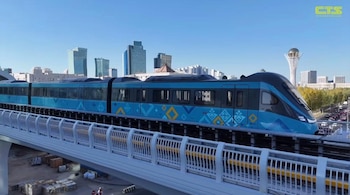 Un tren azul y gris con patrones decorativos transita sobre una vía elevada. Al fondo se ve la ciudad de Astana con edificios modernos y la Torre Baiterek bajo un cielo azul claro