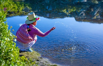 Mujer con sombrero y manta multicolor agachada junto al agua mientras lanza gotas con la mano sobre una laguna