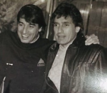 Fotografía en blanco y negro de dos hombres con vestimenta oscura, uno de ellos sonriente y el otro con chaqueta de cuero