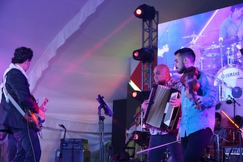 Tres músicos tocan guitarra, acordeón y violín en un escenario iluminado con luces de colores. Detrás se ve una batería Yamaha y una pantalla grande