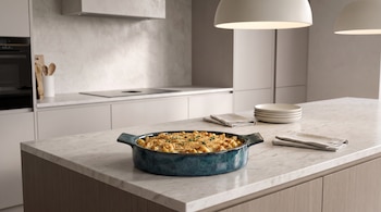 Una fuente azul con pasta gratinada, platos apilados y servilletas sobre una isla de mármol en una cocina.