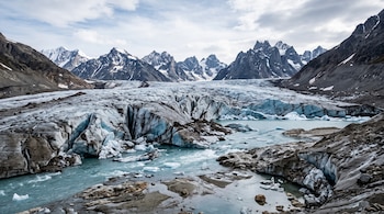 Glaciar con grietas en el hielo, agua de deshielo en primer plano y montañas rocosas al fondo bajo un cielo parcialmente nublado.