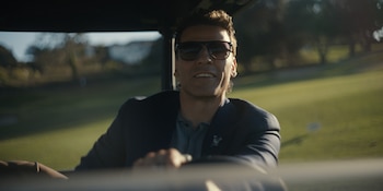 Primer plano de Charles Melton en un carrito de golf, sonriendo, con gafas de sol y una chaqueta oscura. Se ve un campo de golf verde y borroso al fondo