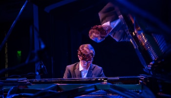 Joven músico con gafas y pelo castaño, vestido de traje, toca un piano de cola oscuro en un escenario con iluminación azul y un reflejo visible
