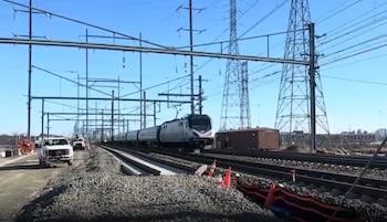 Un tren circula por una vía férrea rodeada de torres eléctricas, catenarias y camionetas de trabajo en un área descubierta