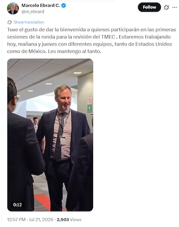 Un hombre de traje y corbata sonríe, lleva un distintivo colgando del cuello, rodeado de otras personas en un pasillo iluminado
