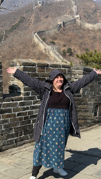 Mujer sonriente con chaqueta de lunares y vestido floral azul, brazos extendidos, de pie sobre la Muralla China. Al fondo, la muralla serpentea por colinas secas