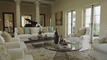 Sala de estar luminosa y elegante con sofás blancos, sillones, dos mesas de centro, un piano de cola y grandes ventanales con vistas a la vegetación