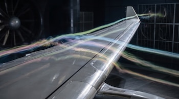 Ala metálica de avión atravesada por líneas luminosas de colores dentro de un túnel de viento con rejillas metálicas al fondo.