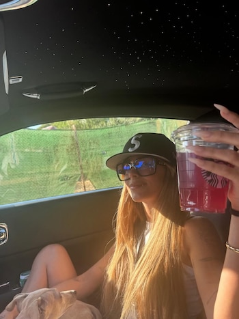 Tini Stoessel sentada en un coche de lujo con techo estrellado. Lleva gorra y gafas de sol, un perro en el regazo y sostiene una bebida rosa