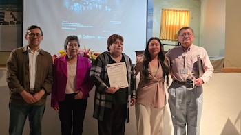 Cinco personas, tres mujeres y dos hombres, posan sonrientes frente a una pantalla de proyección con texto. Flores decoran una mesa frente a ellos