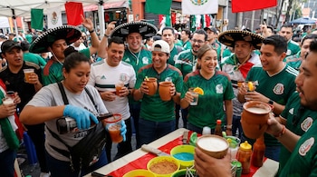 Una multitud de personas con camisetas de fútbol de México y sombreros de charro bebiendo en un puesto callejero con banderas mexicanas.