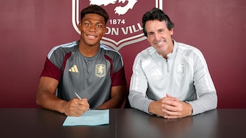 Un futbolista y un entrenador sentados en una mesa, el futbolista firma un documento. Detrás, un muro con el escudo del Aston Villa F.C