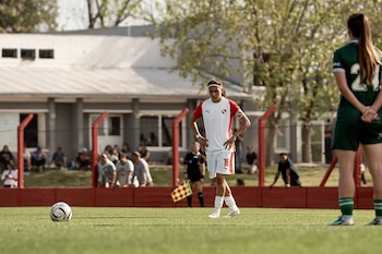 Una jugadora de fútbol con uniforme blanco y rojo permanece de pie cerca del balón en un campo de césped frente a otra jugadora