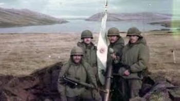 Cuatro soldados con uniformes verdes y cascos posan en una trinchera, con una bandera en un poste. Al fondo, un cuerpo de agua y colinas