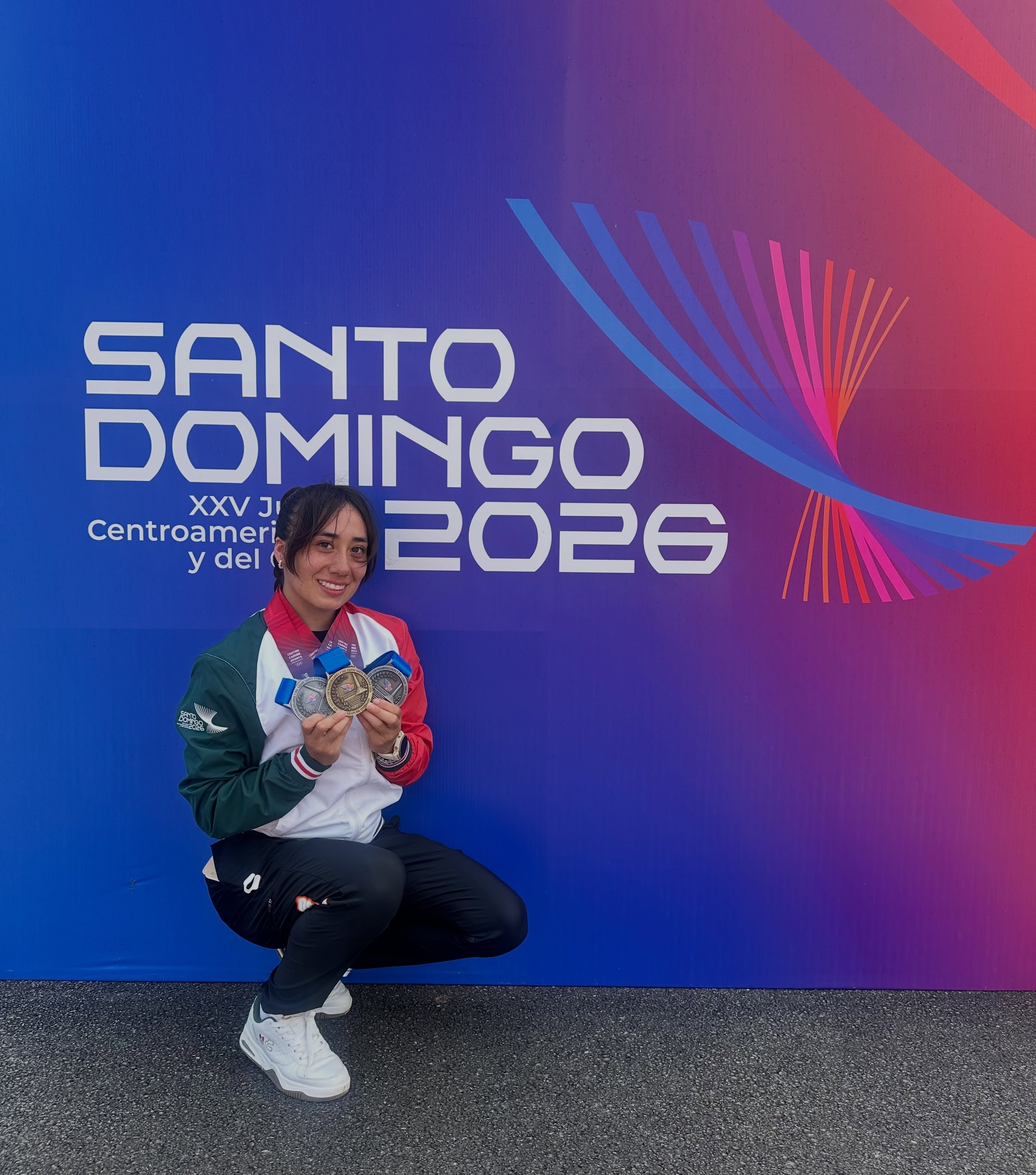 La pentatleta mexicana Catherine Mayran Oliver posa en cuclillas con sus medallas frente al panel promocional de los Juegos Centroamericanos y del Caribe. (Instagram: Catherine mayran)