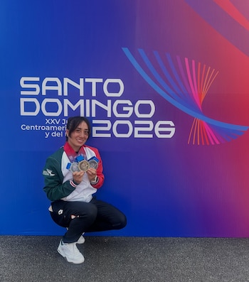 Una atleta agachada sostiene varias medallas frente a un fondo azul y morado con la inscripción Santo Domingo 2026