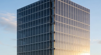 Vista angular de un edificio alto cubierto completamente con paneles solares semitransparentes bajo un cielo azul con nubes ligeras. Una cara está iluminada con luz dorada.