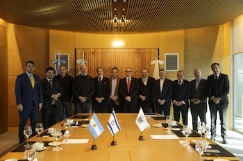 Doce hombres posan de pie en un salón. En primer plano, una mesa de reuniones con banderas de Argentina, Israel y una blanca con escudo, vasos y tazas