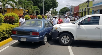 Un automóvil sedán azul y una camioneta pickup blanca con daños menores en una calle. Varias personas, árboles, arbustos y edificios. Matrícula dominicana.