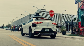 Un coche Waymo blanco con sensores en el techo avanza por una calle. Se ve un cartel de 'STOP ALL WAY' y un edificio con patrón geométrico en el fondo