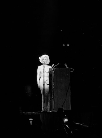 Marilyn Monroe en un escenario oscuro, de pie y sonriendo ante un micrófono con un vestido brillante y ajustado. Un atril de madera se ve a su derecha