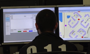Espalda de un hombre sentado frente a dos monitores de computadora que muestran un formulario de datos y un mapa de calles con edificios y marcadores